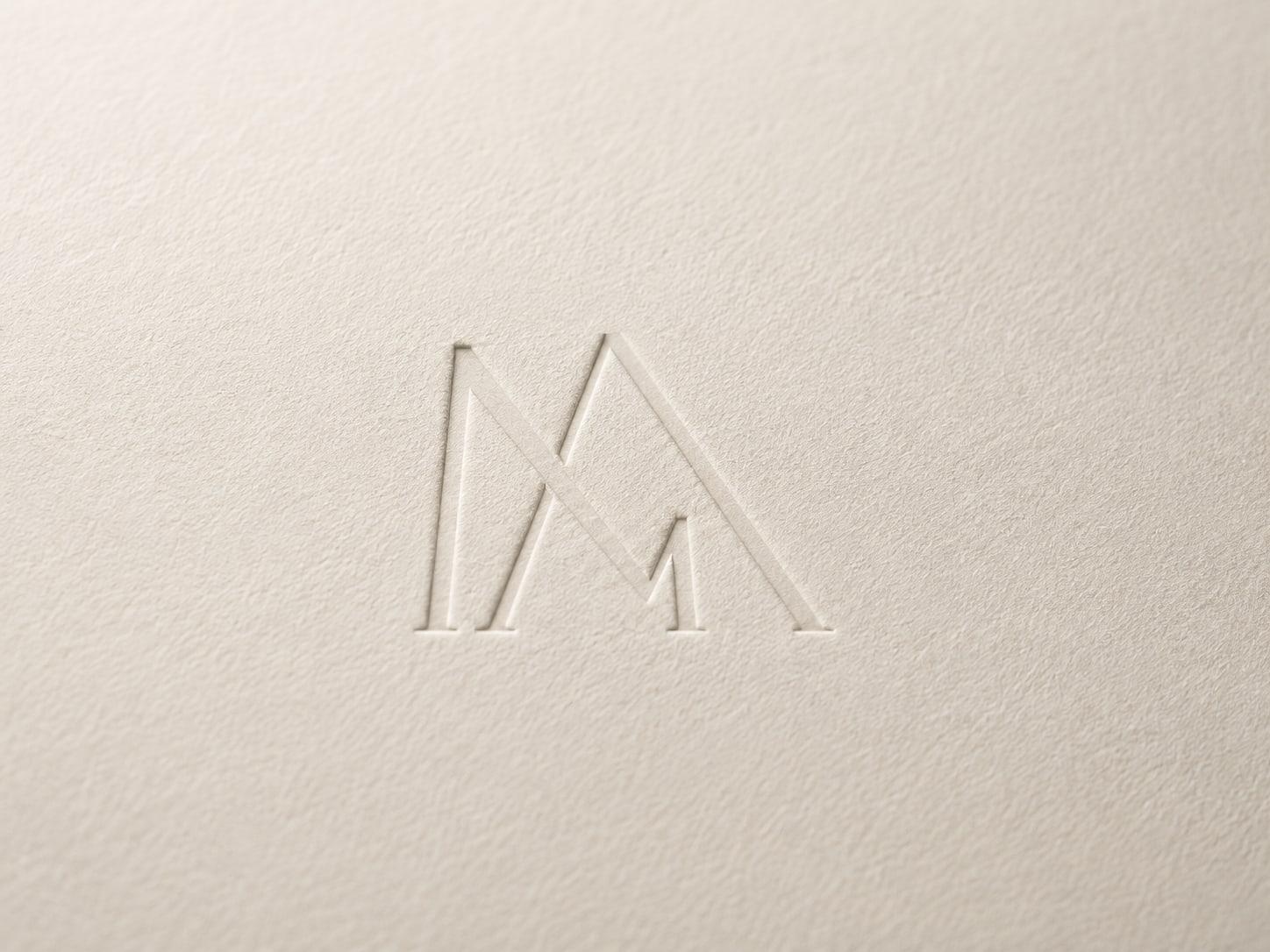 A minimal geometric monogram debossed into thick cream cotton paper, lit at a raking angle so the impression casts a soft shadow.