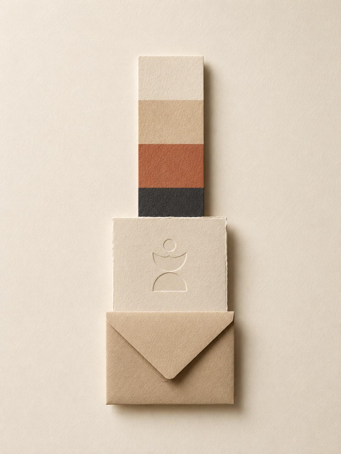 A stationery set on cream paper: a stacked colour swatch strip in sand, terracotta and charcoal above a deckle-edged card embossed with an abstract figure mark, tucked into a kraft envelope.